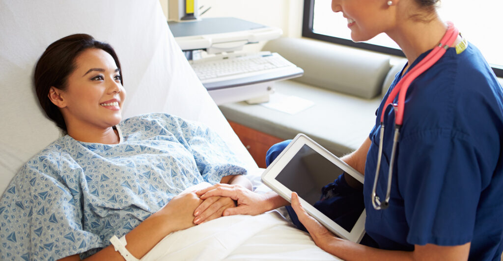 A nurse with a tablet speaking with a patient who is laying in a bed